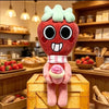 A plush doll resembling a red, slightly monstrous face with large black eyes and white teeth, standing in front of wooden she