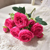 A bouquet of pink silk peony roses with green leaves displayed on white lace fabric.