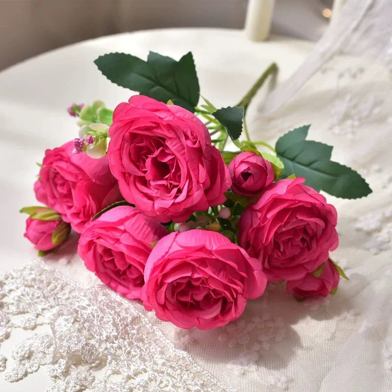 A bouquet of pink silk peony roses with green leaves displayed on white lace fabric.