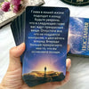 A hand holds a dark blue tarot card with white text and a silhouette of a person standing on a rock against a purple night sk