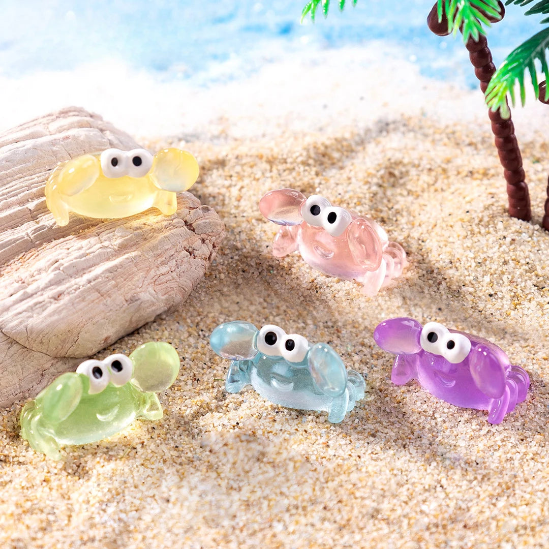 A set of luminous resin mini crab decor figures in yellow, pink, and purple resting on a sandy beach.
