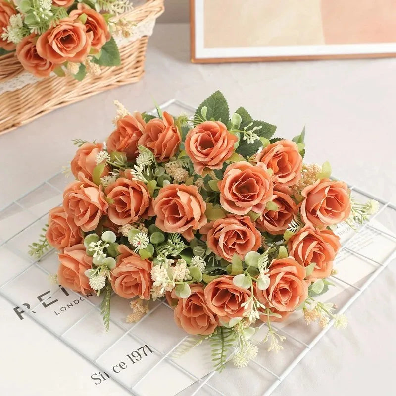 A bouquet of five silk touch artificial orange roses with green foliage arranged on a white surface.