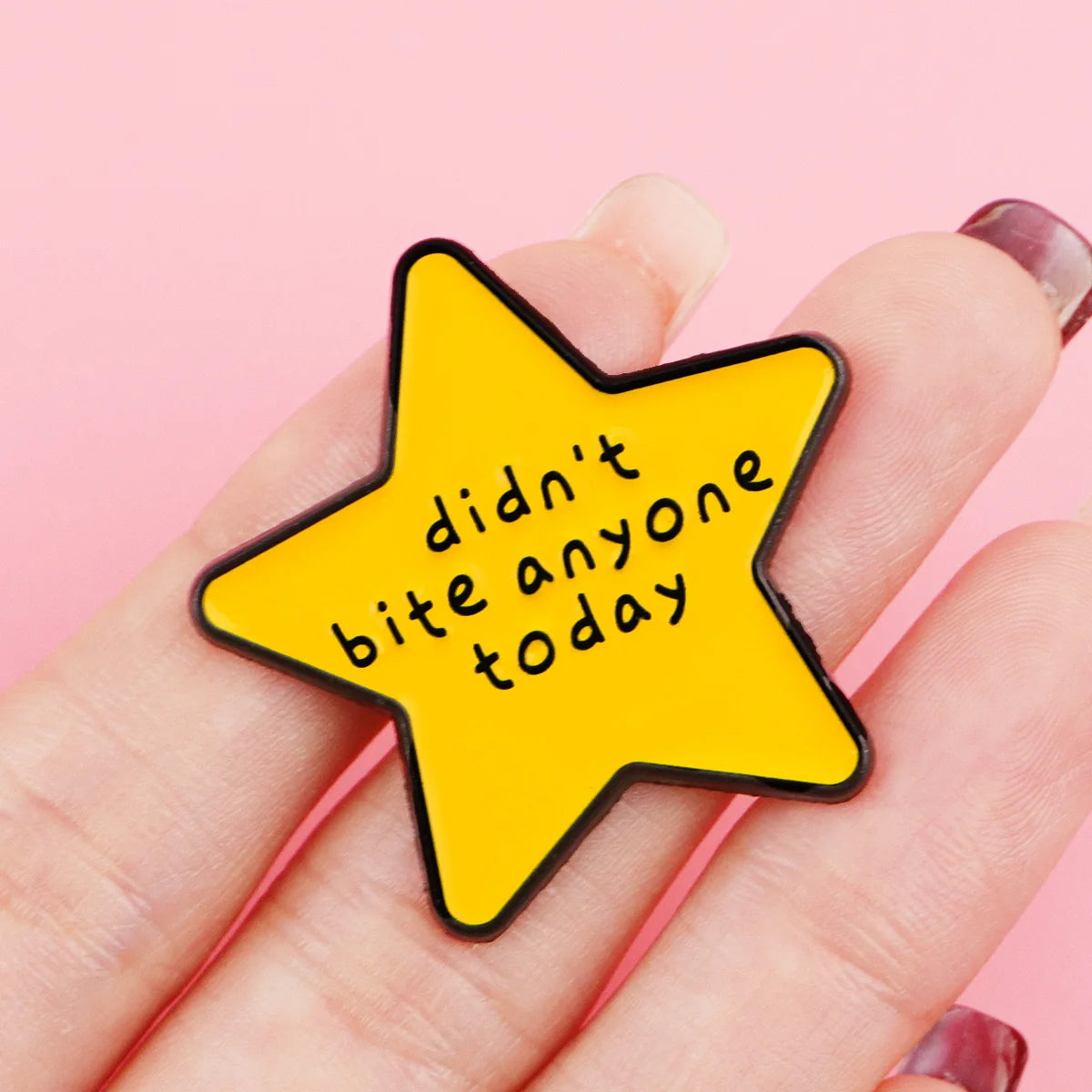 A hand holds a yellow star enamel pin with the text didn't bite anyone today on it.