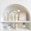 A minimalist ceramic vase is displayed on a white shelf with dried greenery and white floral elements.