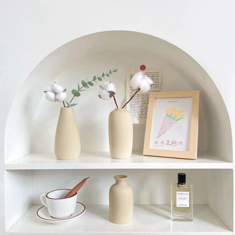 A minimalist ceramic vase is displayed on a white shelf with dried greenery and white floral elements.