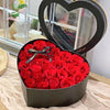 A bouquet of red roses in a heart-shaped gift box with a clear lid and black ribbon.