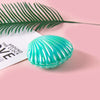 A turquoise seashell product resting on a pink surface with a green palm leaf in the background.