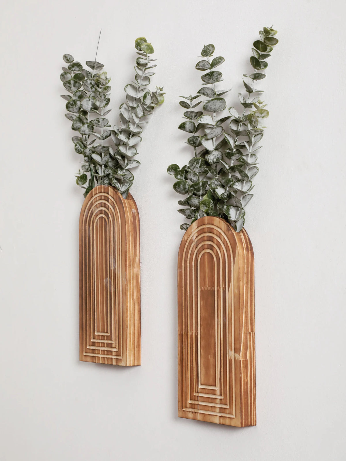Un par de jarrones de madera arqueados con follaje verde contra un fondo blanco.