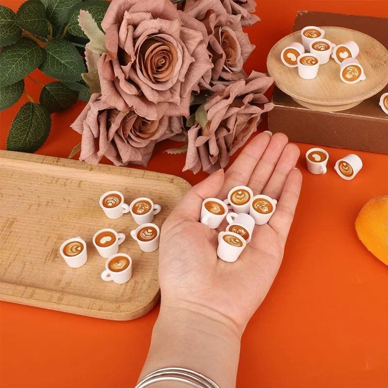 A hand holds ten miniature white resin coffee mugs on an orange background next to dried roses and a wooden board.