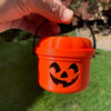 An orange miniature pumpkin Halloween bucket with a carved face is held by a hand against a green background.