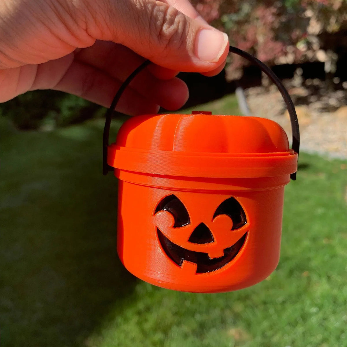 An orange miniature pumpkin Halloween bucket with a carved face is held by a hand against a green background.