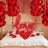 A display of red heart-shaped balloons and rose petals decorating a bed for a romantic celebration.