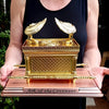 A golden metal statue of the Ark of the Covenant resting on a pink base.