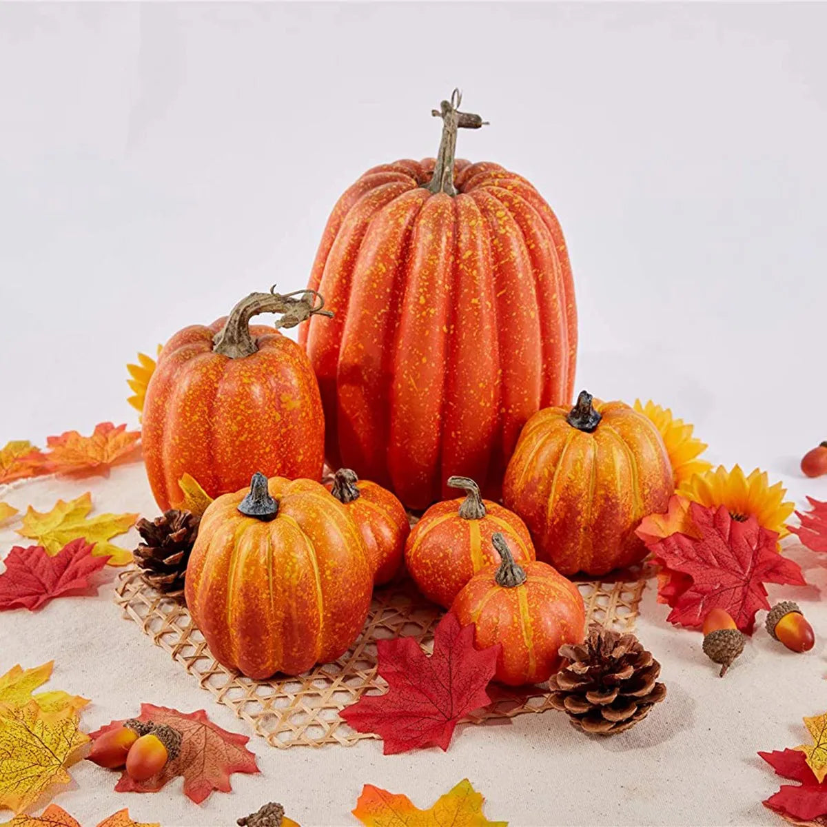 A set of seven orange artificial pumpkins arranged on a light surface with autumn leaves and pinecones.