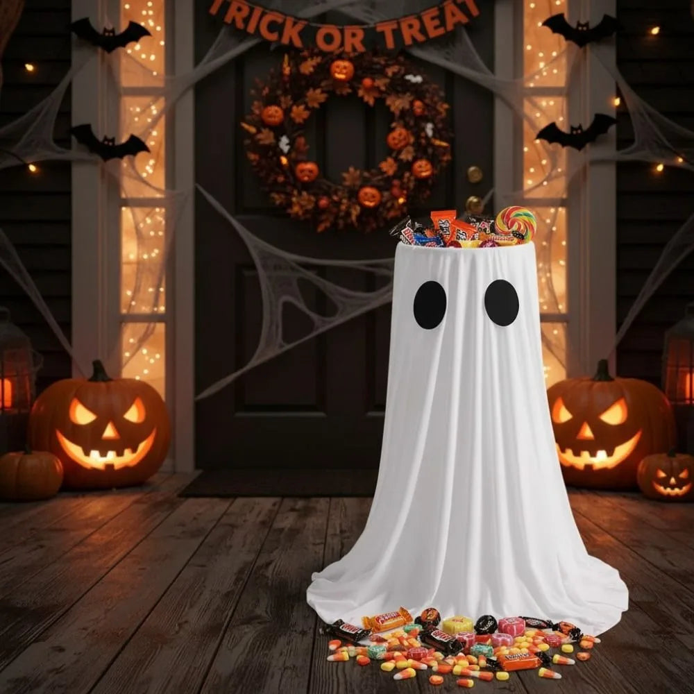 A white ghost-shaped fabric display with black eyes sits on a wooden floor in a Halloween decorated setting.