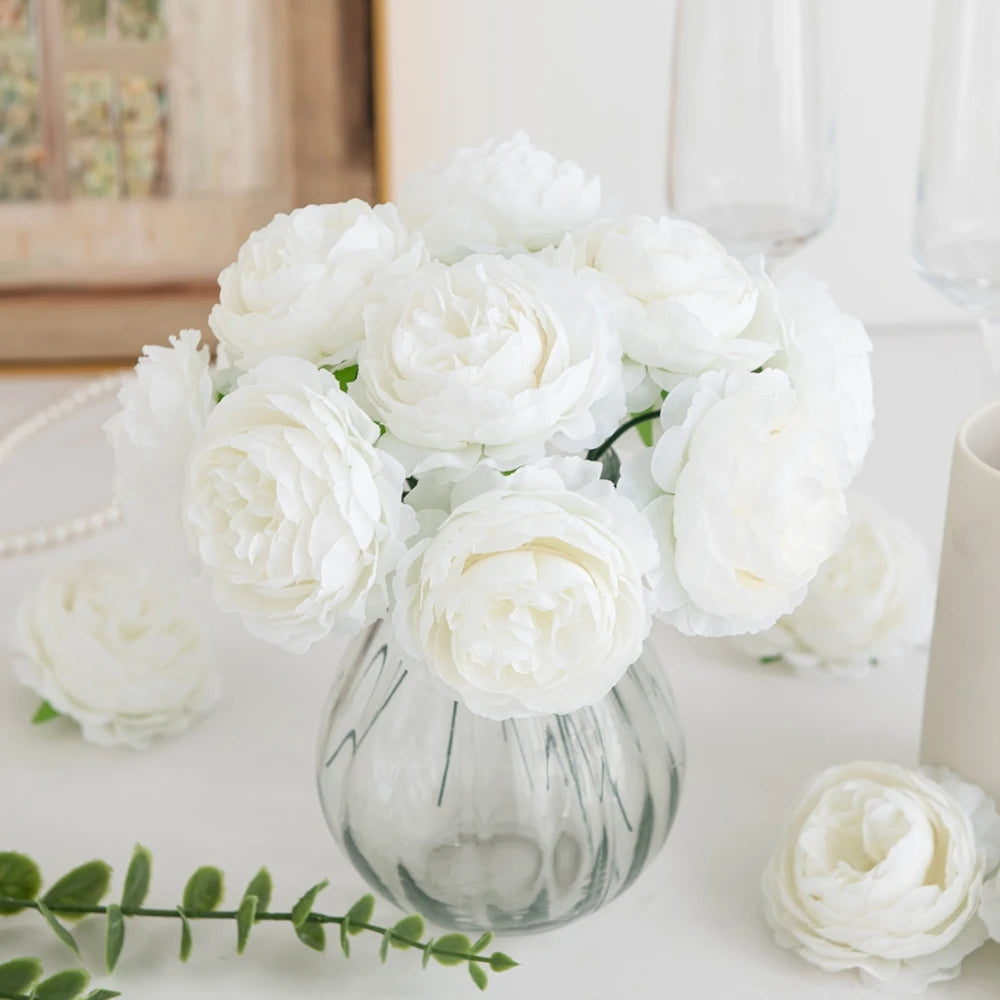 A bouquet of white silk peony artificial flowers in a clear glass vase on a white surface.