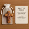 A handcrafted wooden prayer cross is displayed inside a white mesh bag on a wooden surface.