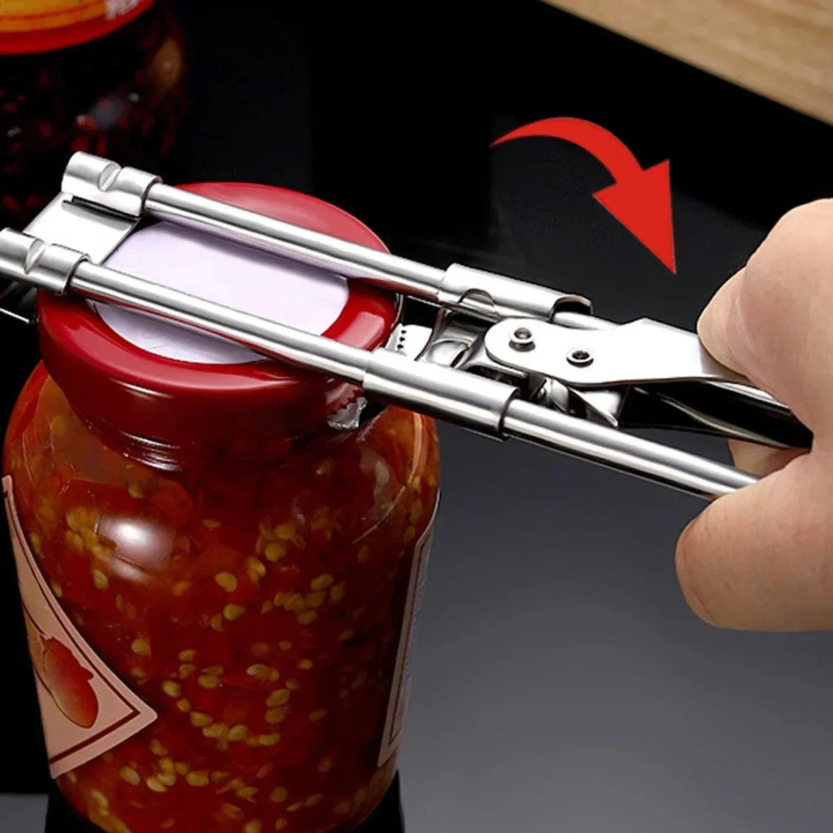 A hand uses an adjustable stainless steel jar and bottle opener to open a red-colored glass jar.