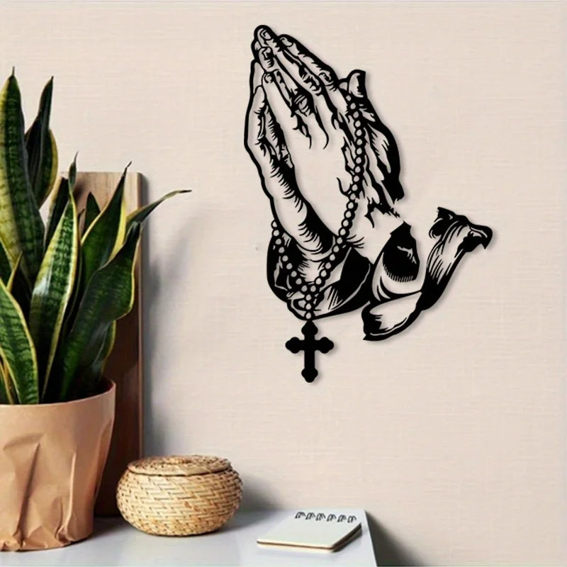 Black metal wall art depicting praying hands with a cross, displayed next to a potted plant and a woven basket.