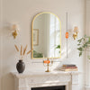 An arched wall mirror with an aluminum frame hangs above a marble fireplace mantel.