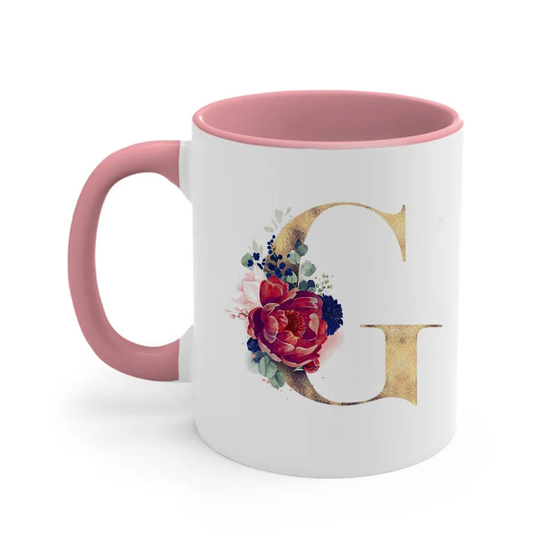 A pink ceramic coffee mug featuring a large gold letter G decorated with floral accents.