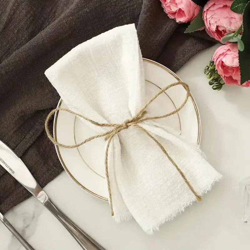 A set of ten white fringed cotton napkins tied with twine rests on a plate next to a dark gray cloth.