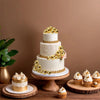 A tiered white cake decorated with gold ball toppers and a pink floral accent, displayed on a wooden table.