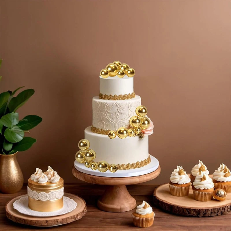 A tiered white cake decorated with gold ball toppers and a pink floral accent, displayed on a wooden table.