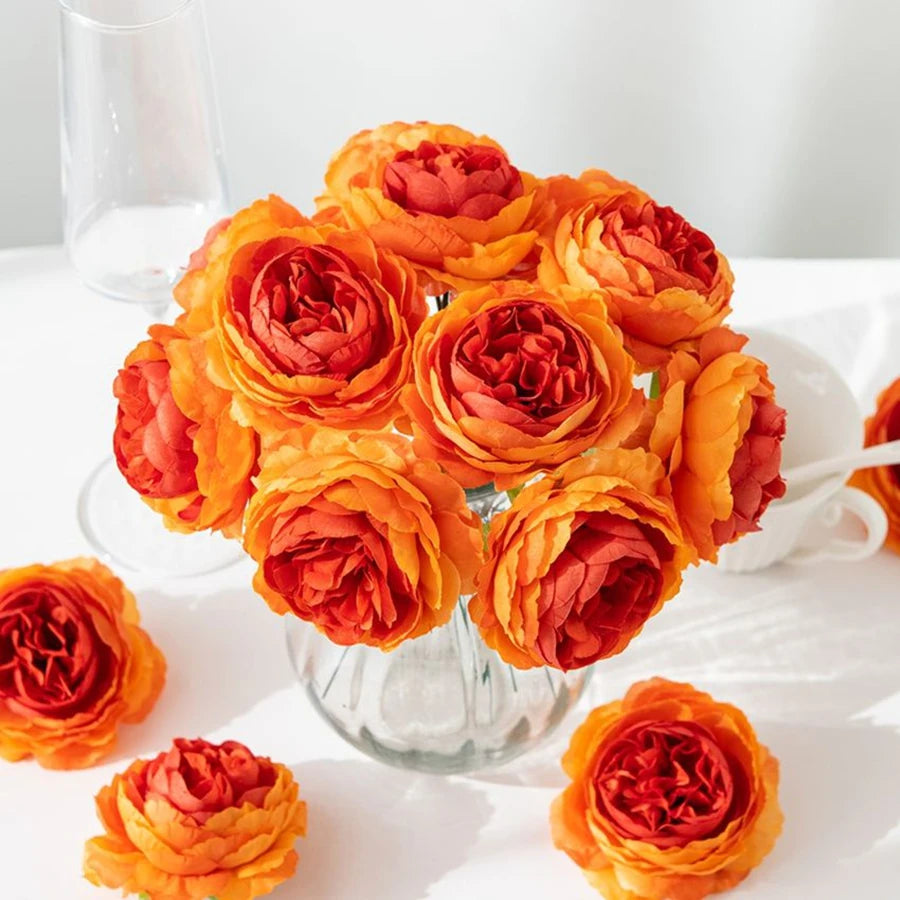 A bouquet of vibrant orange and red silk peony artificial flowers in a glass vase on a white surface.