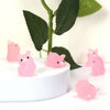 Pink glow-in-the-dark mini pig figurines displayed on a white surface with green leaves in the background.