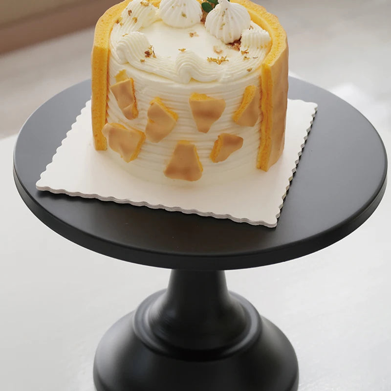 A white layer cake decorated with cream and gold accents displayed on a black metal cake stand.