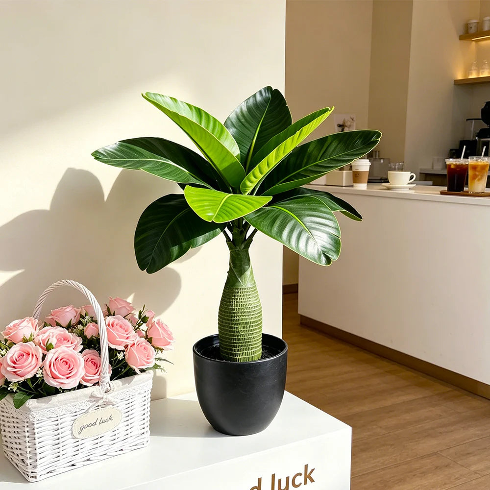 Un árbol de plátano artificial en maceta con grandes hojas verdes sobre una superficie blanca junto a una cesta tejida.
