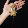 A hand holds a wooden Our Lady of Guadalupe pendant charm with a gold-colored oval frame and a dark tassel.