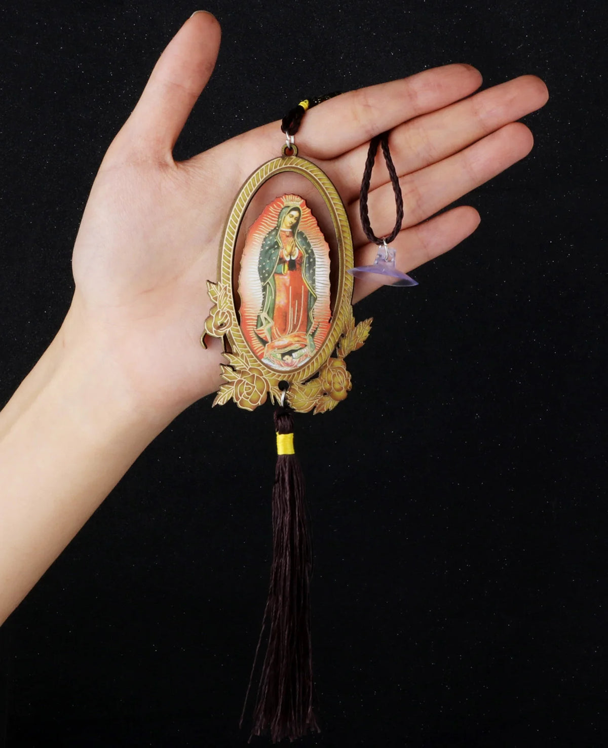 A hand holds a wooden Our Lady of Guadalupe pendant charm with a gold-colored oval frame and a dark tassel.