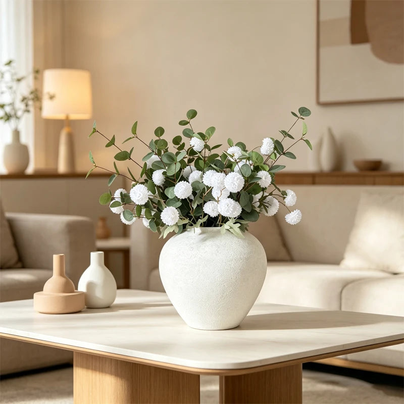 A white textured round vase filled with green foliage sits on a light wood table in a modern living room setting.