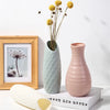 A light green ceramic vase holds dried yellow flowers on a white surface next to a framed picture and a wooden object.