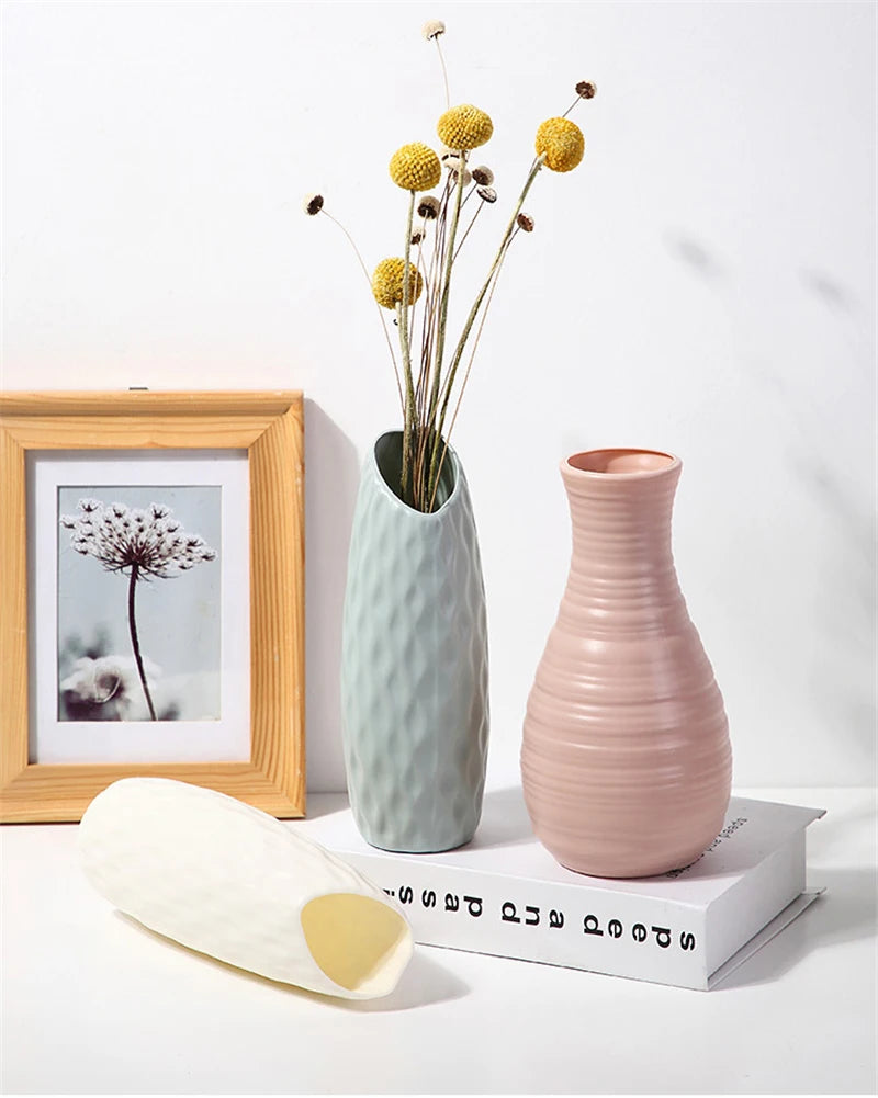 A light green ceramic vase holds dried yellow flowers on a white surface next to a framed picture and a wooden object.