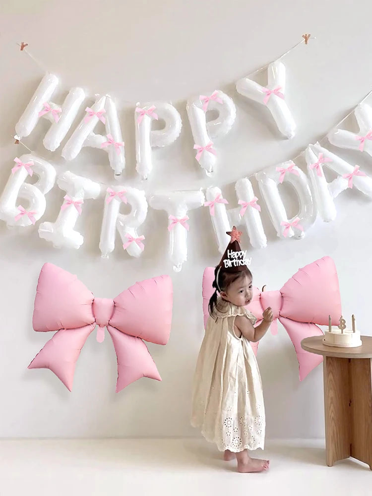 A young girl stands in front of a white balloon banner spelling out Happy Birthday, surrounded by pink and white balloons.
