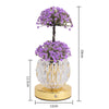 A small potted purple flower tree in a clear glass vase with gold accents, sitting on a round gold base.
