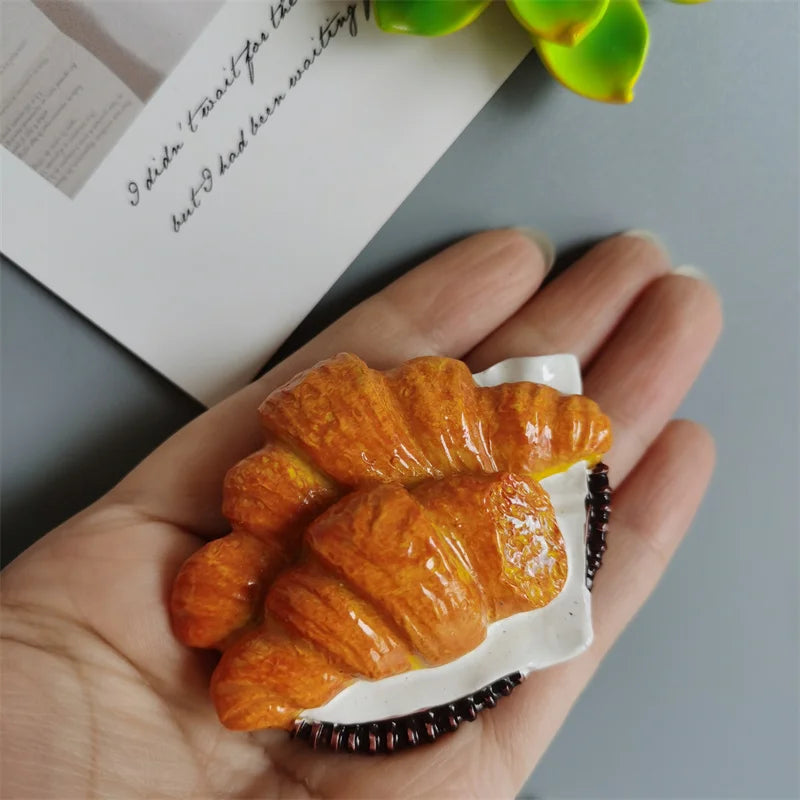 A golden croissant food-shaped fridge magnet resting on a white card against a gray background.