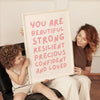 A woman holds a framed canvas with the text You Are Beautiful Strong Resilient Precious Confident And Loved in front of a chi