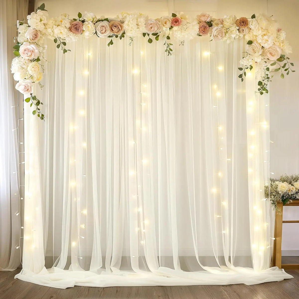 A sheer white chiffon backdrop curtain decorated with floral arrangements and string lights for an event or wedding.