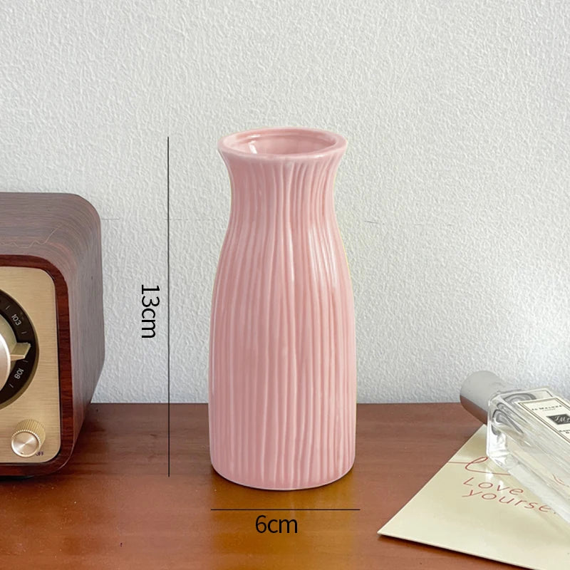 A tall, pink ceramic bud vase with vertical ribbing sits on a wooden surface next to a vintage-style appliance.
