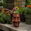 A terracotta planter shaped like a smiling face holds succulents and flowers on a wooden surface.