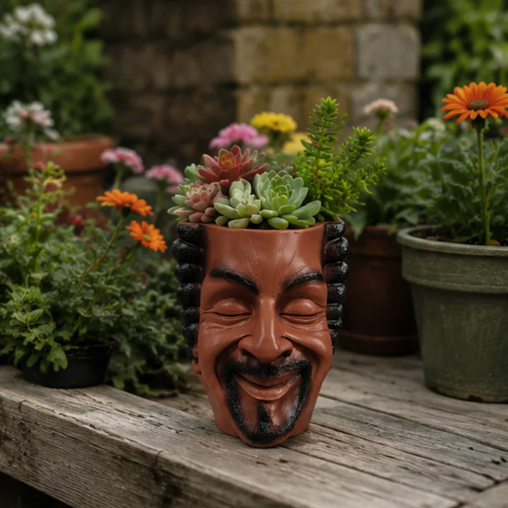 A terracotta planter shaped like a smiling face holds succulents and flowers on a wooden surface.
