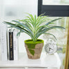 A potted artificial chlorophytum branch with green leaves sits on a white surface next to books and an alarm clock.