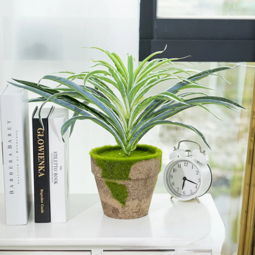 A potted artificial chlorophytum branch with green leaves sits on a white surface next to books and an alarm clock.