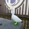 A white garden statue of a goose with a yellow beak and green feet sitting on a gray stone ledge.