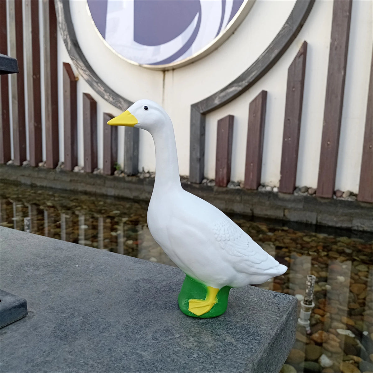 A white garden statue of a goose with a yellow beak and green feet sitting on a gray stone ledge.