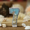A glitter number cake topper shaped like the number 7 with a starfish design, set against a blurred background of white sea s
